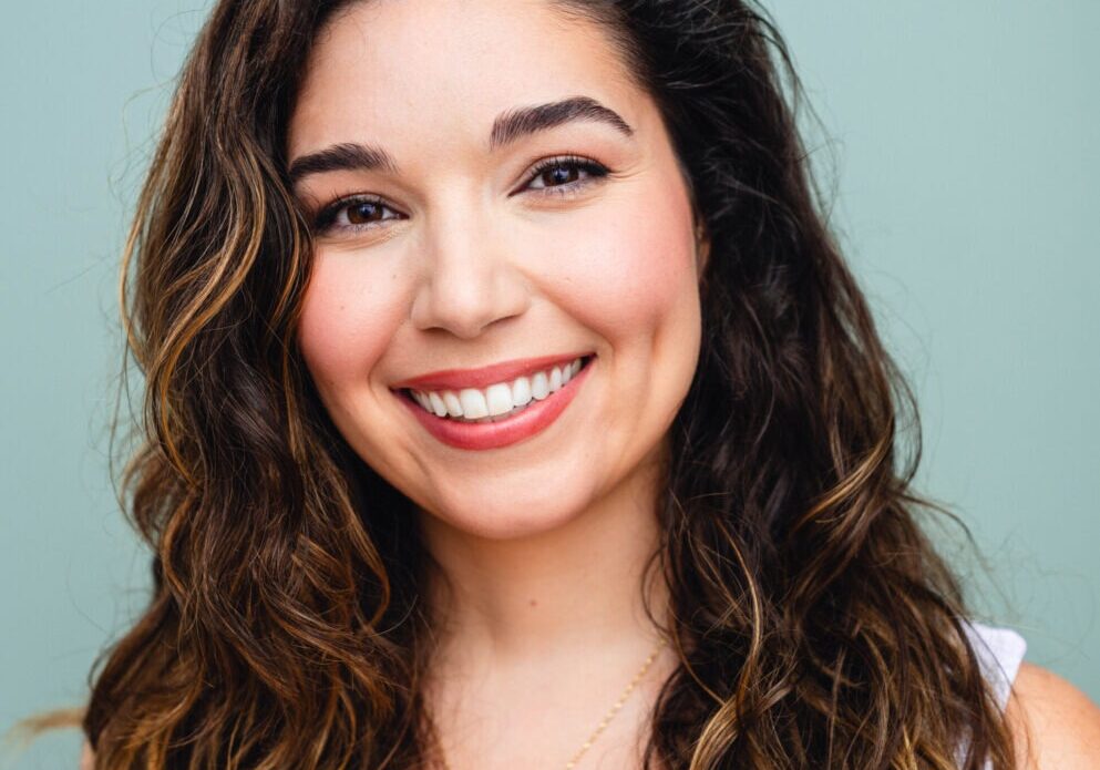 headshot of female voiceover coach Emma Romasco wearing white top and standing in front of of a light blue green background
